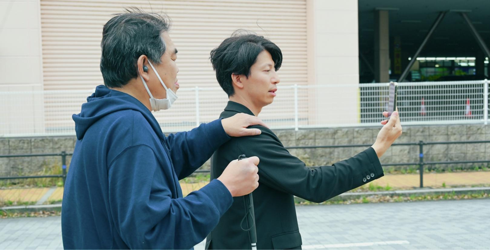 髙田将平さんがスマートフォンを掲げ白杖を持った男性に肩を貸している