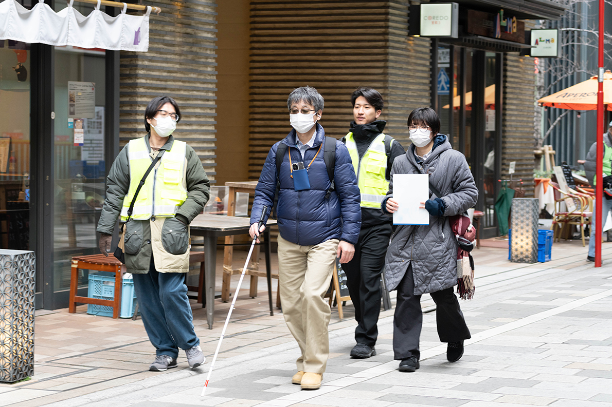 街の中の歩道を、白杖を持つ人を含む複数人が並んで歩いている様子