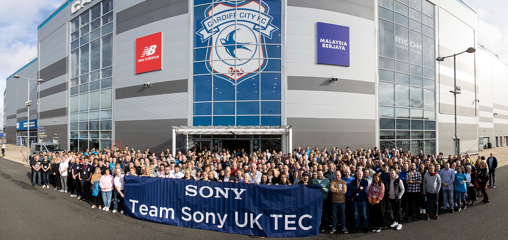 建物の前に大勢の参加者が集合しており、"Team Sony UK TEC"の文字が記載されている青い横断幕を持っている