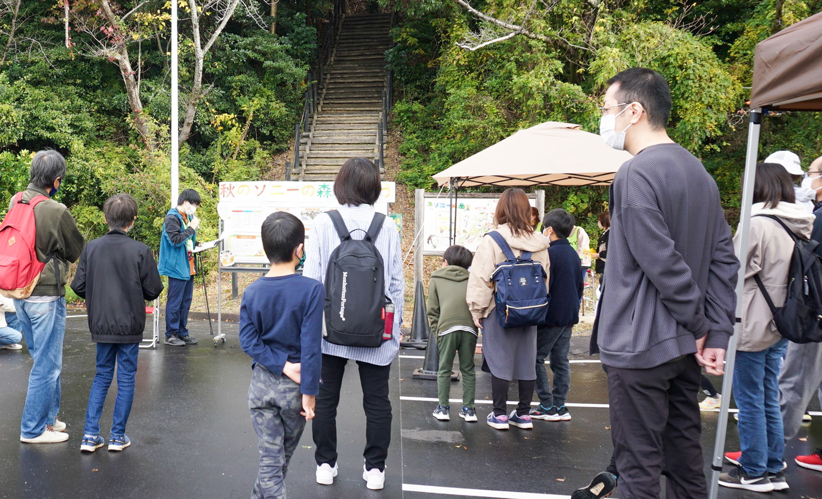 丘の公園の入り口に集まるたくさんの親子連れ。公園の入口は豊かな緑に囲まれ、木の階段が見える