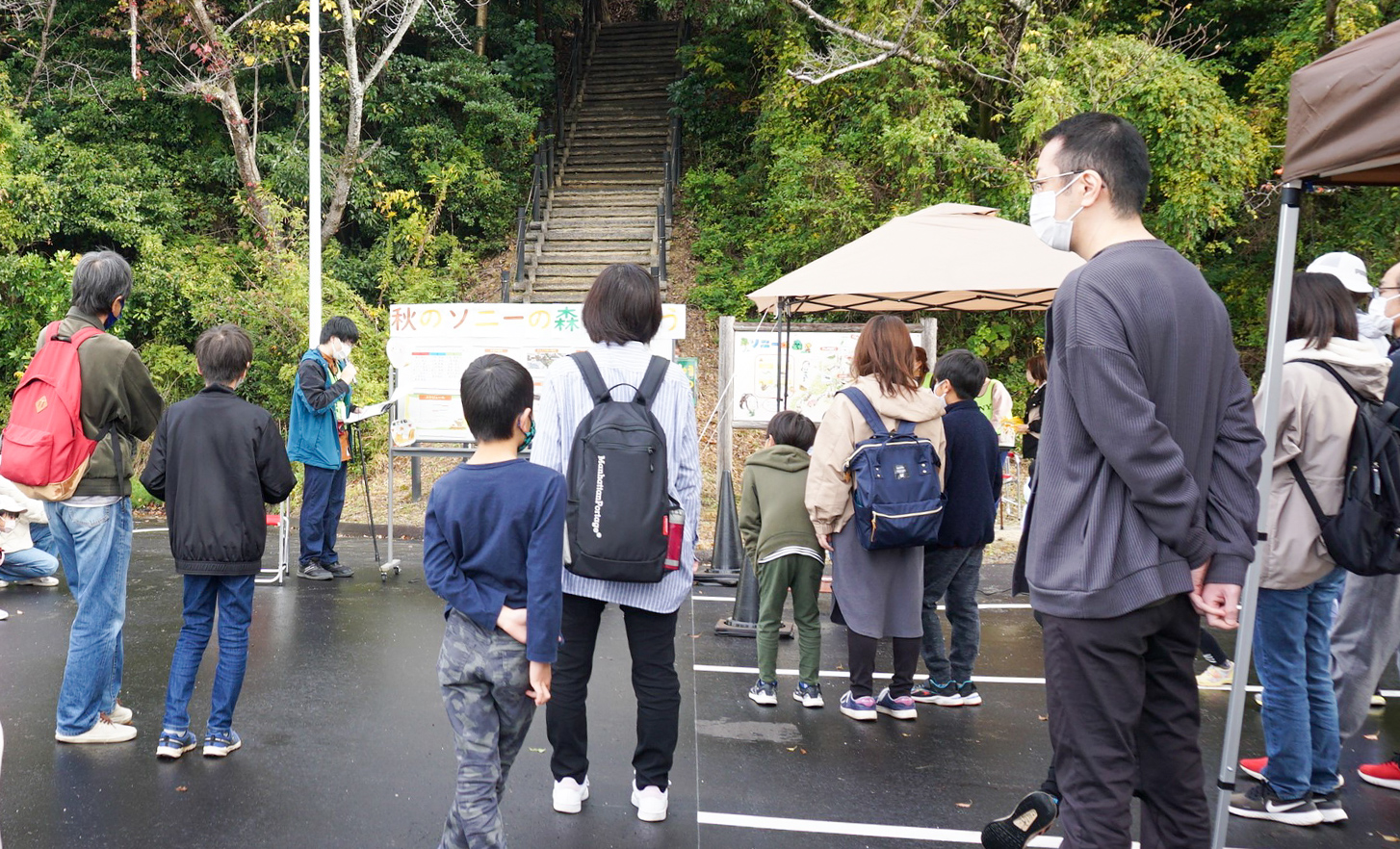 丘の公園の入り口に集まるたくさんの親子連れ。公園の入口は豊かな緑に囲まれ、木の階段が見える