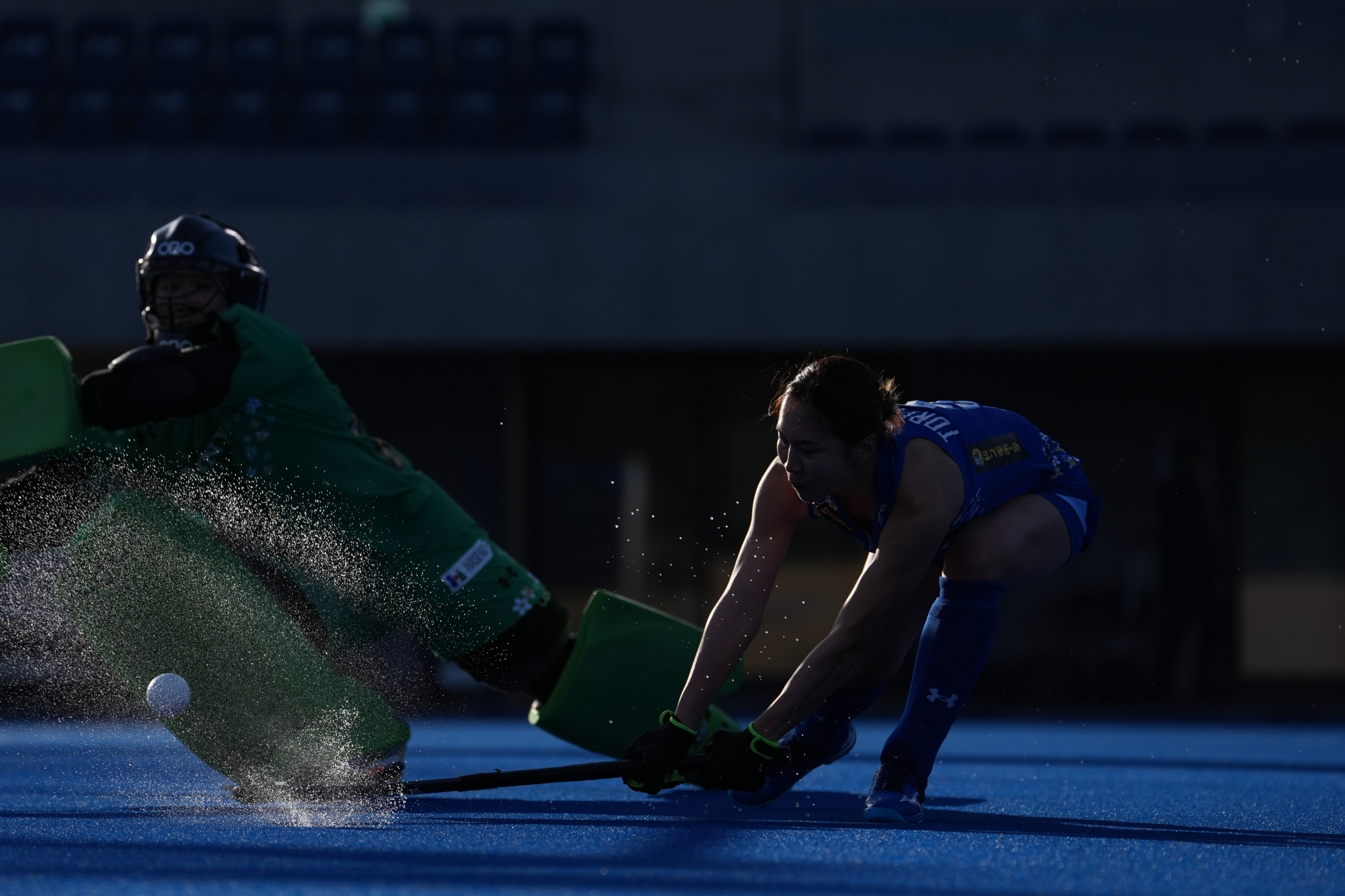 Field hockey player taking a shot against a goalie on a blue pitch. Click to open modal