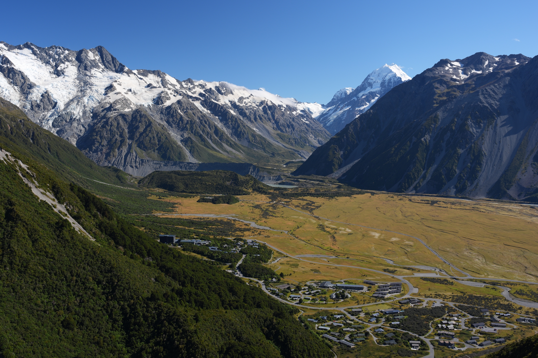 A landscape photo of a wide valley and grasslands surrounded by snow-capped mountains Click to open modal