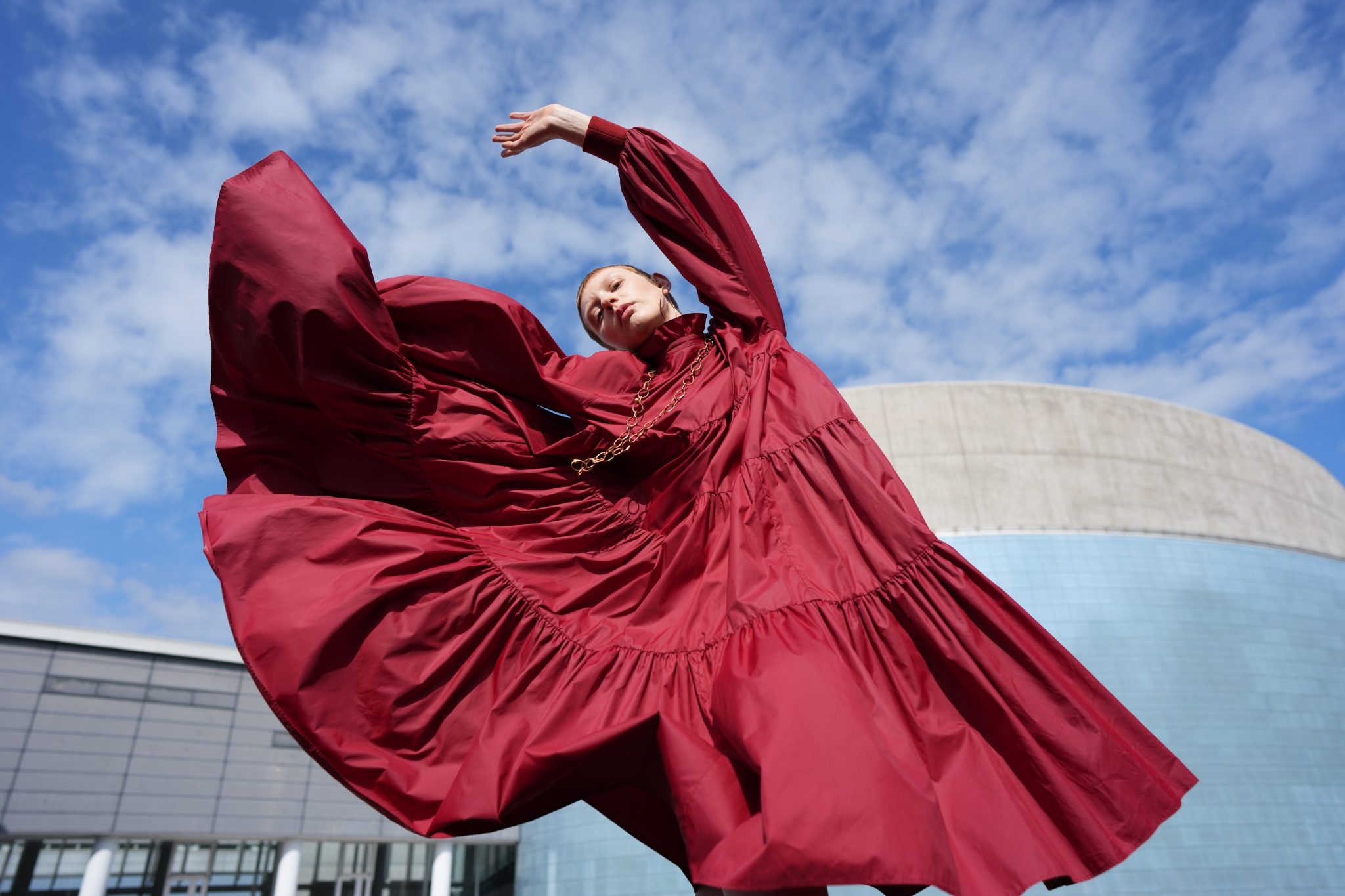 A portrait of a woman jumping with her red dress billowing widely under a blue sky Click to open modal