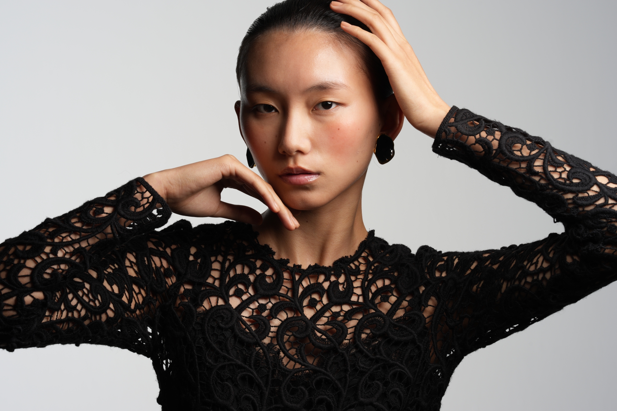 A portrait of a woman wearing a black lace long-sleeve top with her hand resting on her head Click to open modal