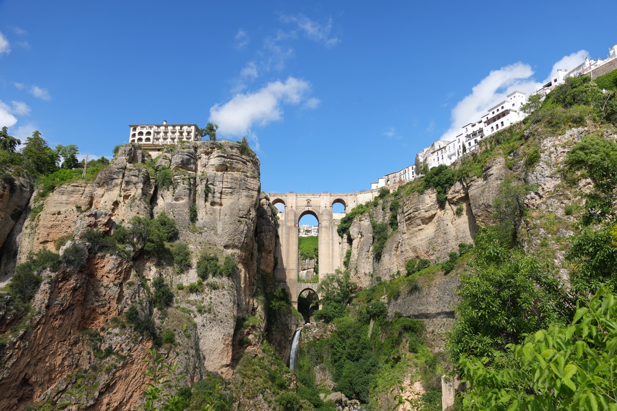 A sample image of the "Puente Nuevo". A stone arch bridge spans a deep, rocky canyon, connecting whitewashed buildings perched precariously atop sheer cliffs under a bright blue sky with scattered clouds. Click to open modal