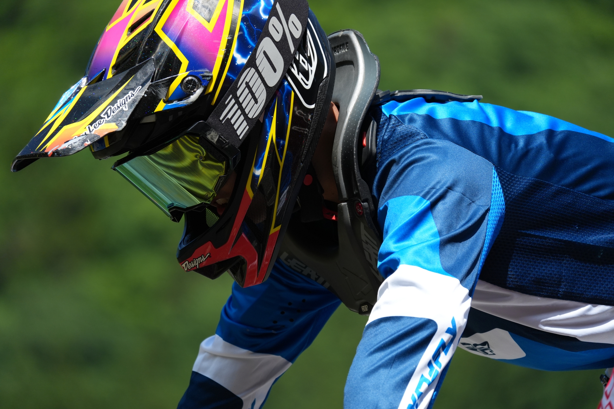 A motocross rider in a colorful helmet and blue gear, leaning forward Click to open modal