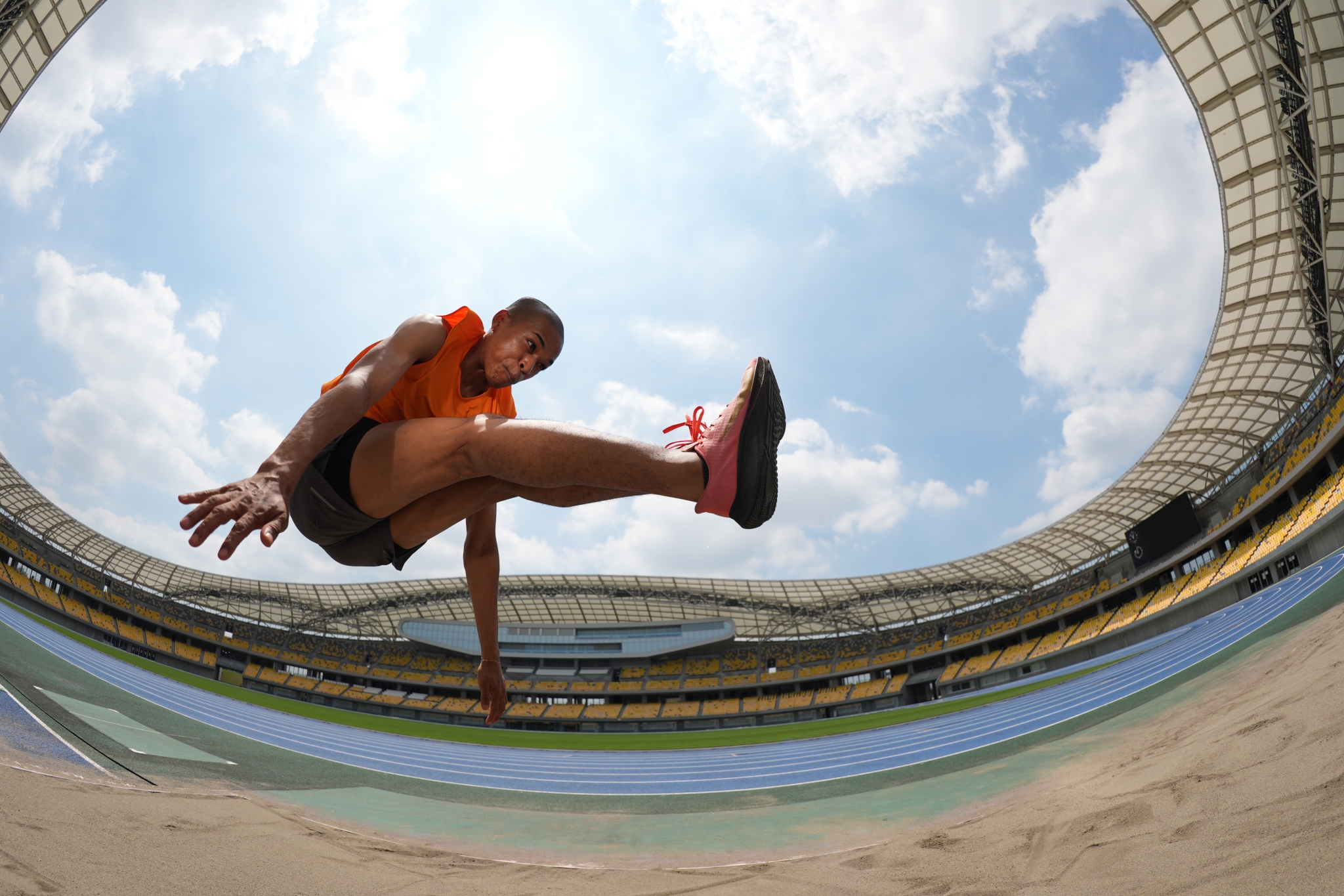 Athlete performing long jump photographed from below with fisheye lens against blue sky and sandy ground Click to open modal