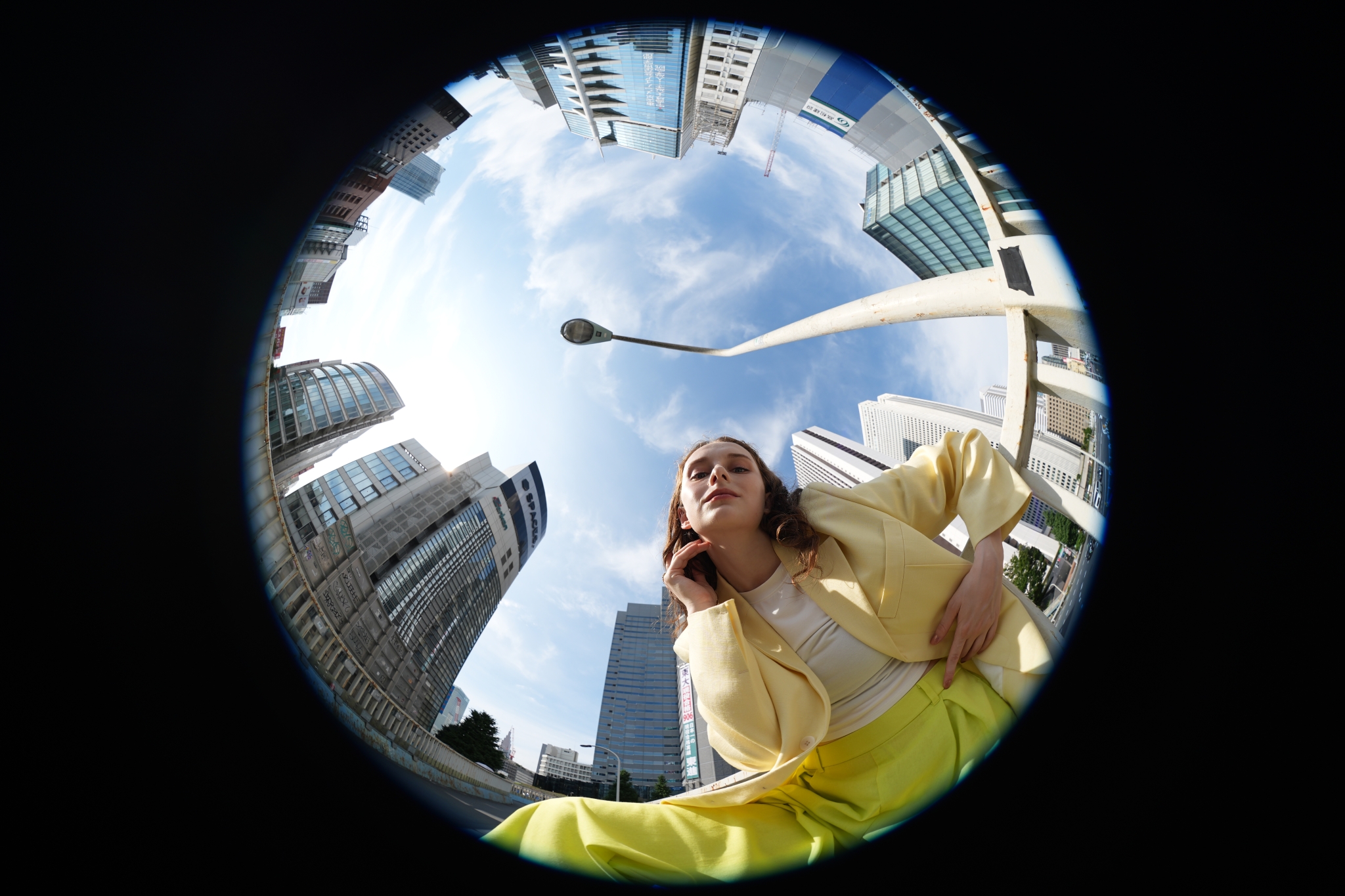Fisheye view looking upward at tall buildings surrounding a person standing against blue sky Click to open modal