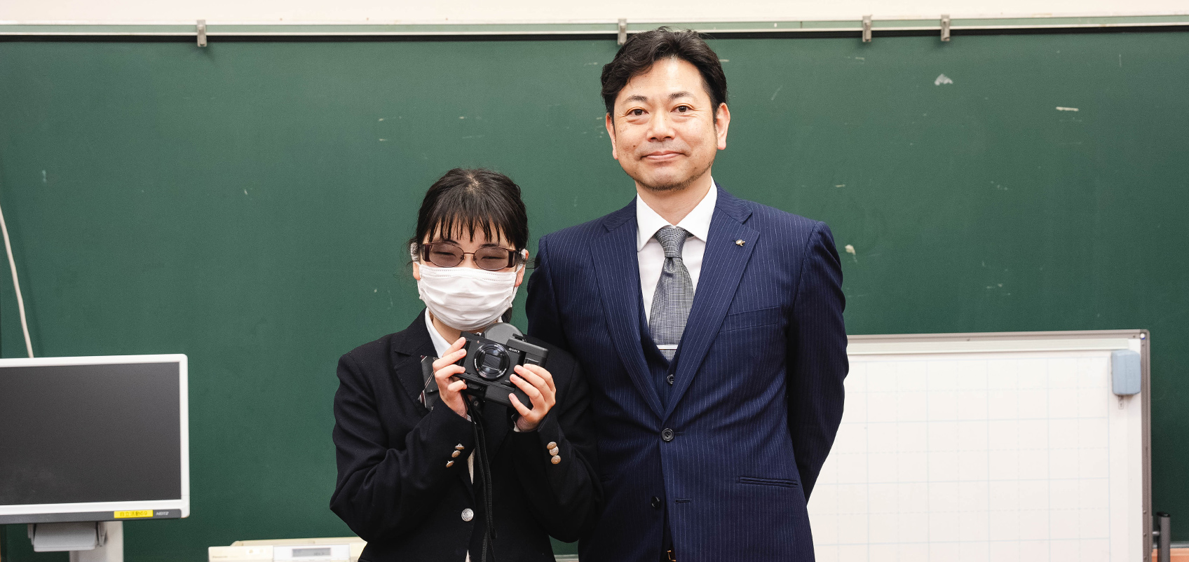 Image taken in a school classroom with blackboard, with a man in a suit and a girl in school uniform smiling for the picture. The girl is wearing a mask, and holding an HX99 RNV retina projection camera kit