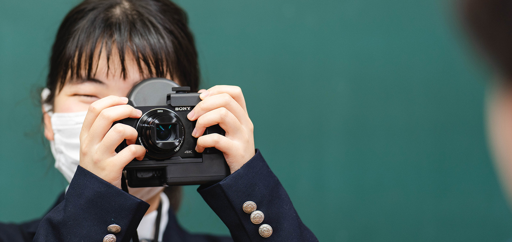 Image taken in a school classroom with blackboard, showing a girl in school uniform, who is wearing a mask and holding an HX99 RNV retina projection camera kit up to her eye, pointing the camera at somebody else who is almost out of shot