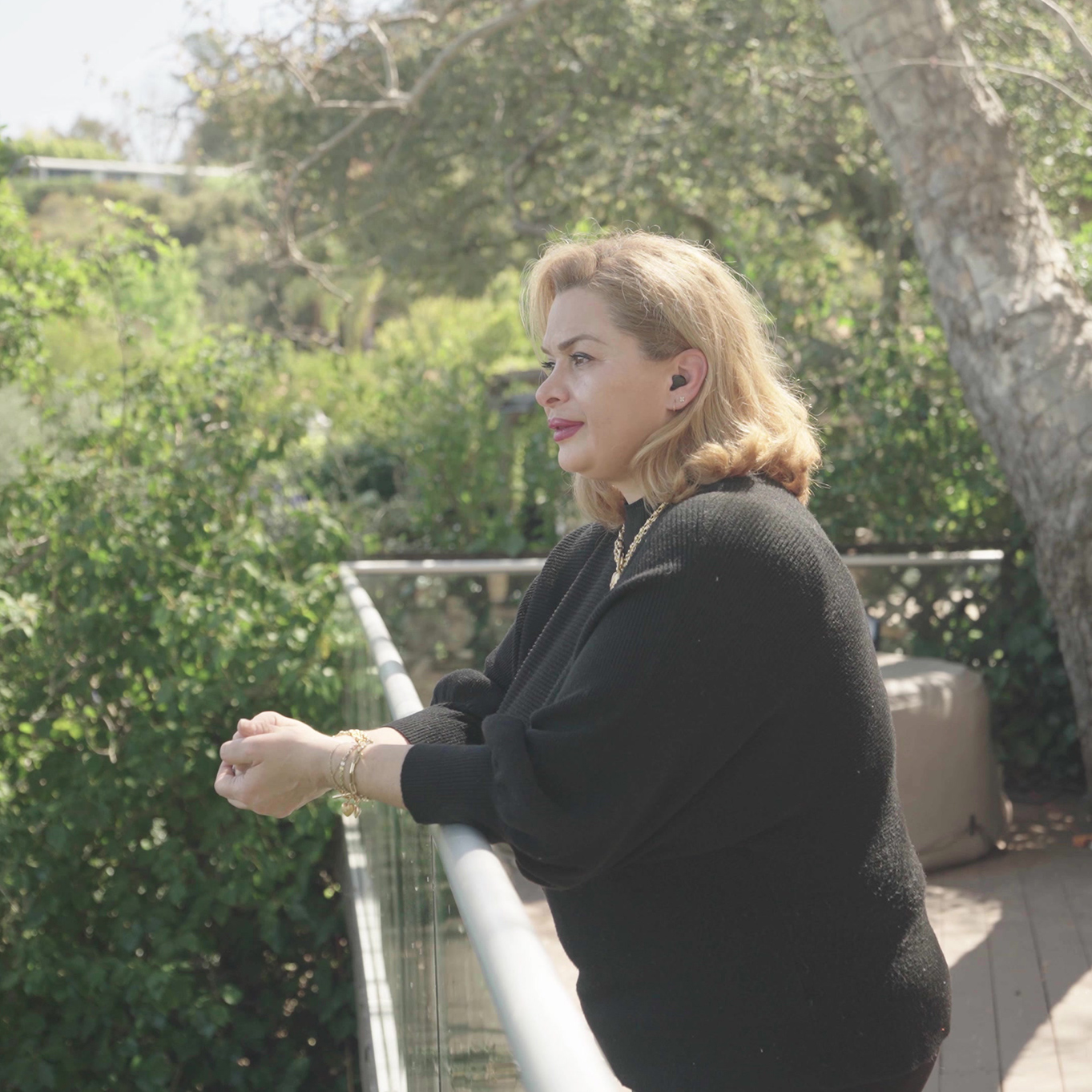 A woman wearing a hearing aid looks out from a balcony surrounded by trees in the warm sun.