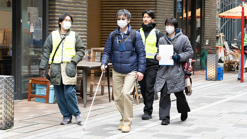 A group of people walking along a city sidewalk, including a person with a white cane.